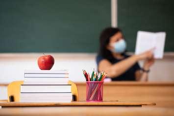 Back to school concept. apple, accessories and books in the classroom, teacher wearing mask in the background. © Can Stock Photo / erika8213
